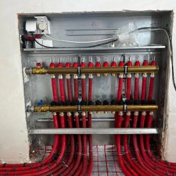 TERMAG SPÓŁKA Z OGRANICZONĄ ODPOWIEDZIALNOŚCIĄ - Rozdzielacz ogrzewania podłogowego z czerwonymi rurami w metalowej szafce, widoczne zawory i przyłącza, instalacja w trakcie montażu w surowym wnętrzu.
