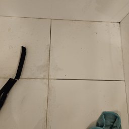 Tomasz Łuszczek - Brudne, białe płytki podłogowe z widocznymi zabrudzeniami w fugach, fragment mopa i szmatki. Widok z góry, ukazujący stan podłogi przed sprzątaniem.