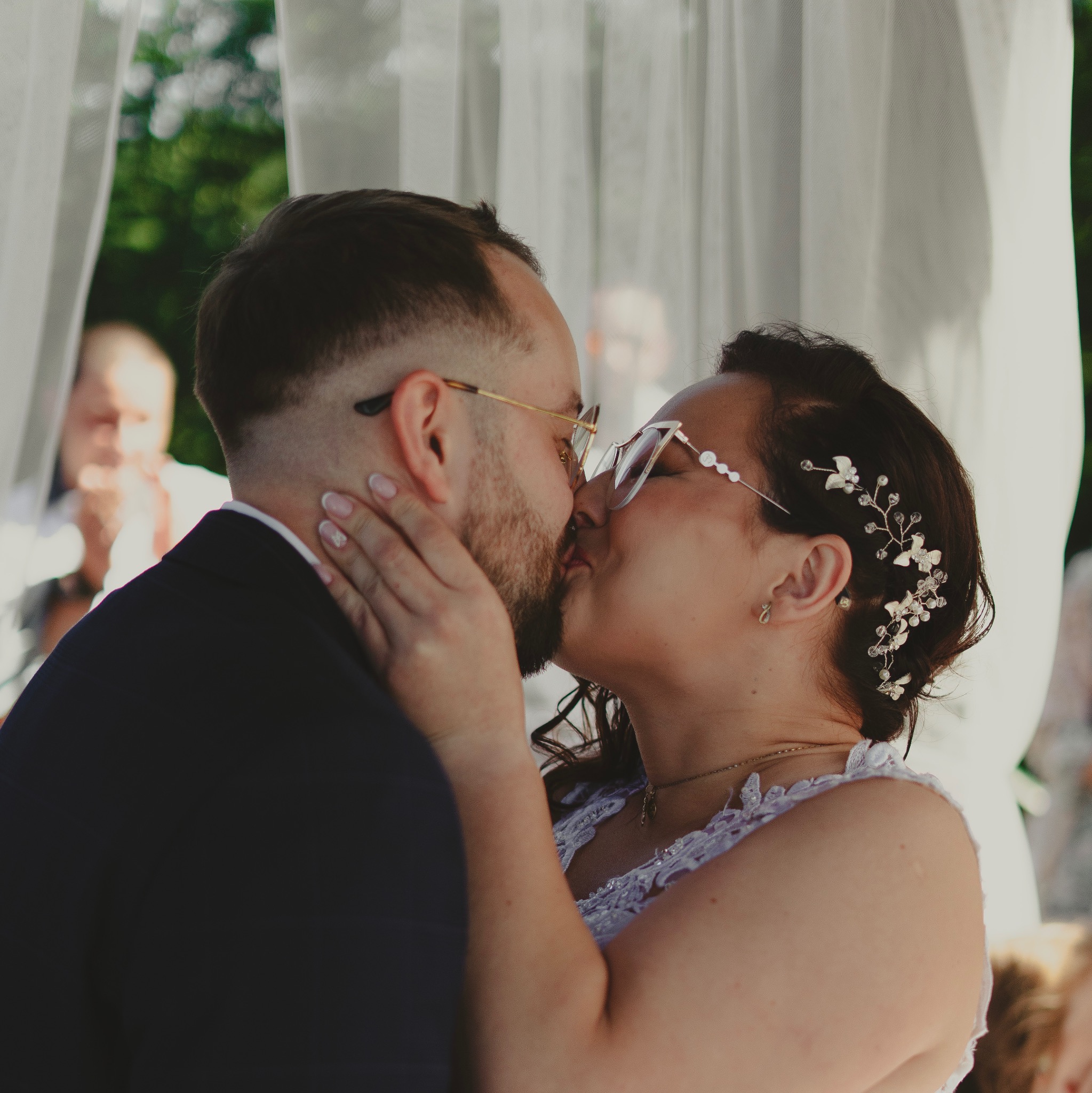Para młoda w okularach, pocałunek pod baldachimem z białych zasłon, panna młoda z ozdobą we włosach.
