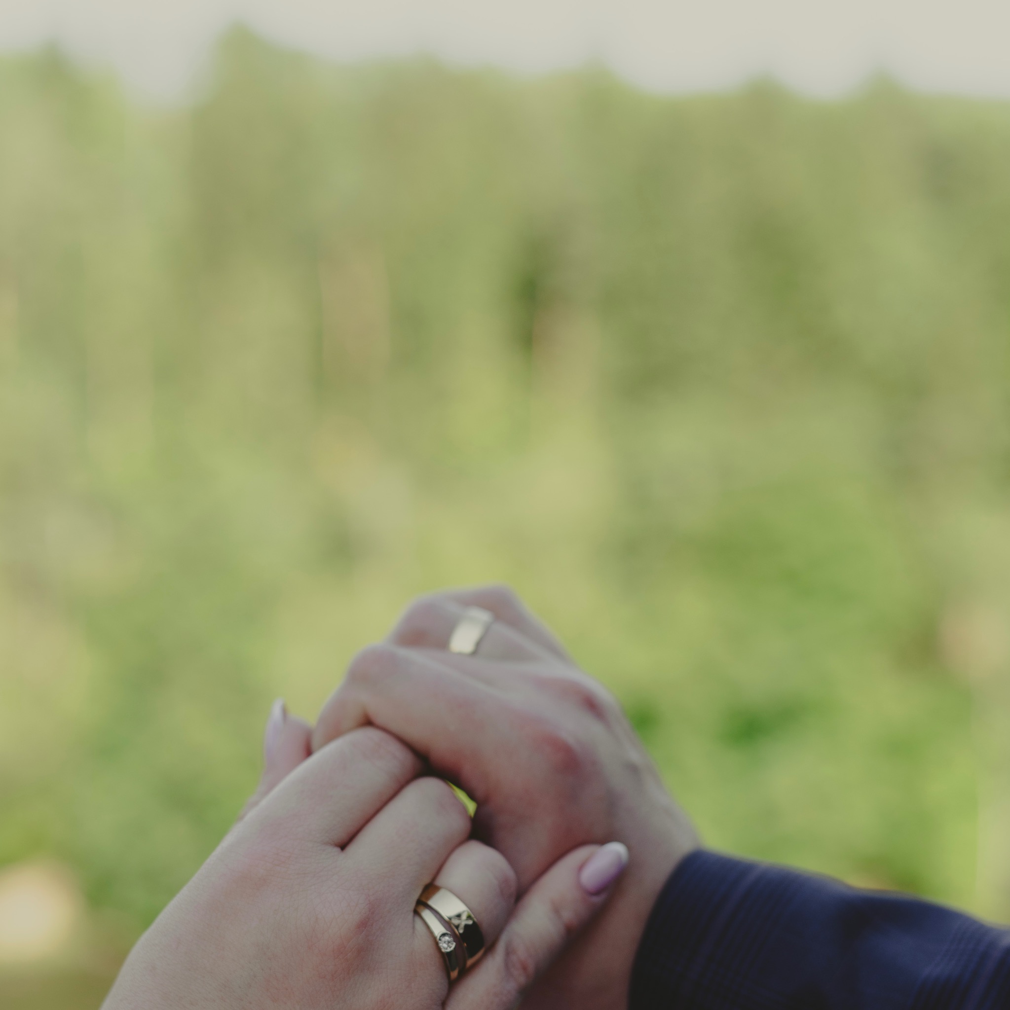 Złączone dłonie pary z widocznymi złotymi obrączkami na tle rozmytej zieleni.