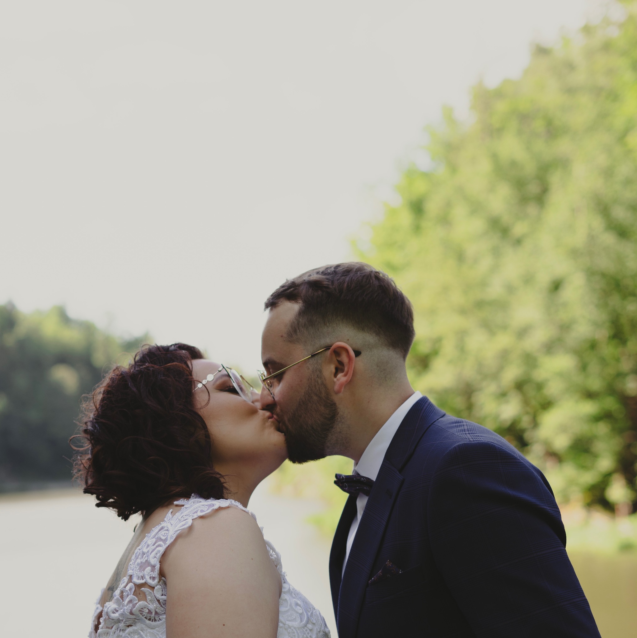 Para młoda w trakcie pocałunku na tle zieleni i wody, panna młoda w koronkowej sukni i ozdobnych okularach, pan młody w granatowym garniturze z muchą.