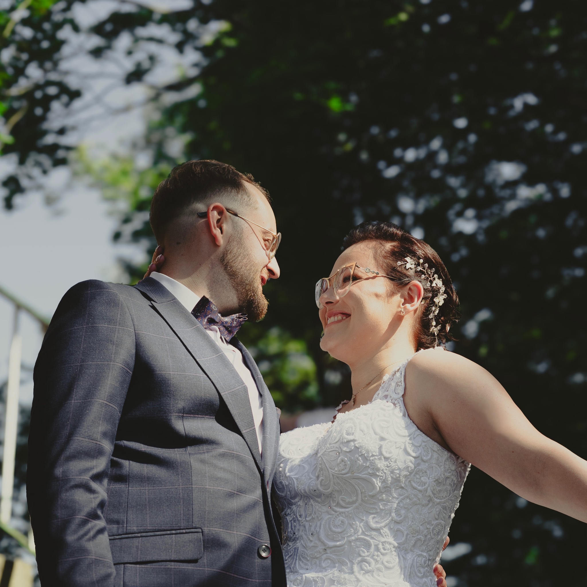 Uśmiechnięta para młoda w okularach, on w kraciastej marynarce i muszce, ona w koronkowej sukni ślubnej i ozdobie we włosach, patrząca na siebie z bliska na tle zieleni.