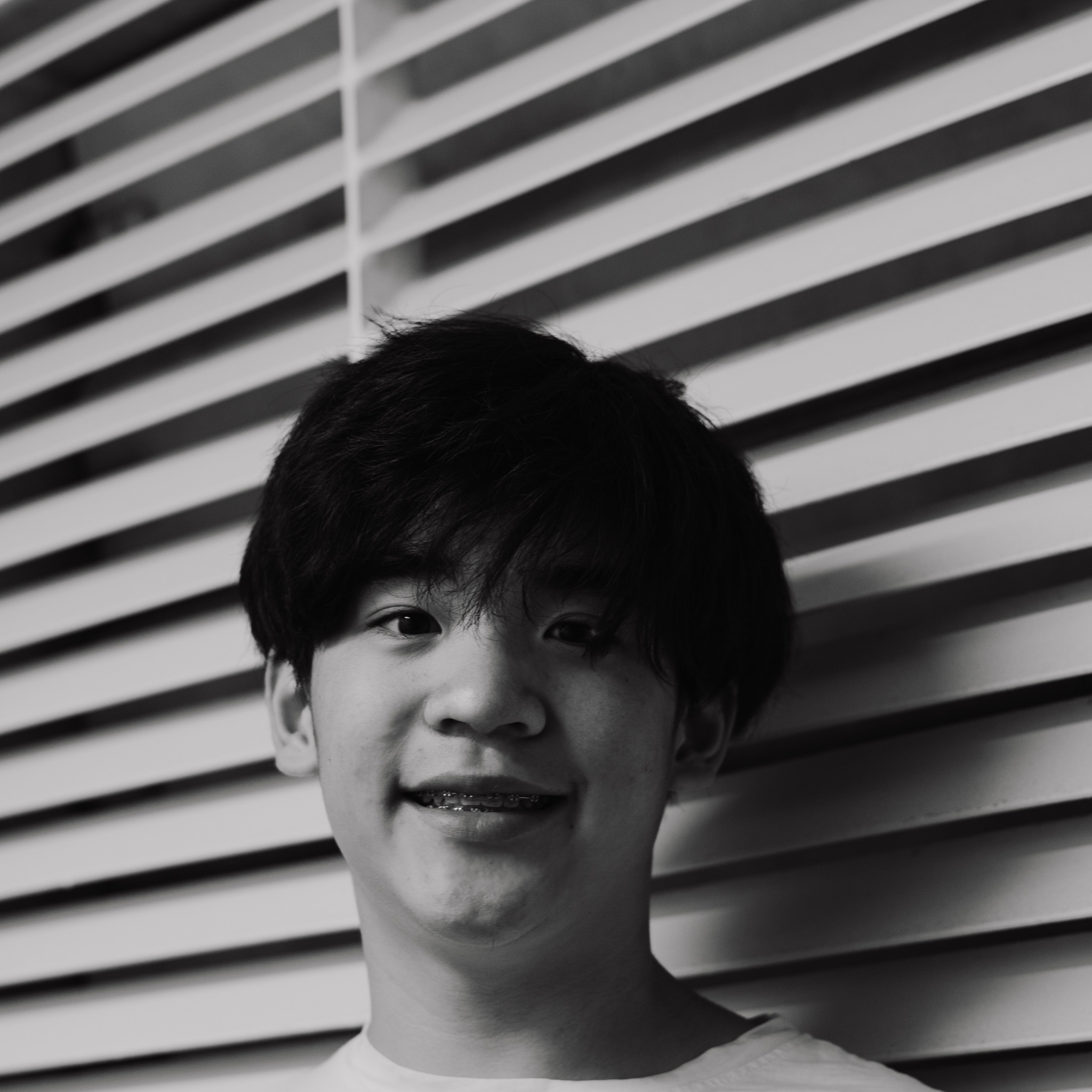 Czarno-biały portret uśmiechniętego młodego mężczyzny z aparatem ortodontycznym na tle żaluzji.