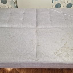 Odnowa i Błysk - Szara rozkładana kanapa z widocznymi plamami przed czyszczeniem, widok z góry na siedzisko z guzikami, drewniana podłoga w tle.