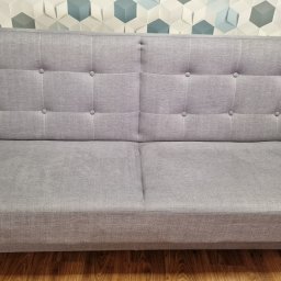 Odnowa i Błysk - Szara, dwuosobowa kanapa z tapicerowanymi guzikami na oparciu, ustawiona na drewnianej podłodze z dekoracyjną ścianą w tle.