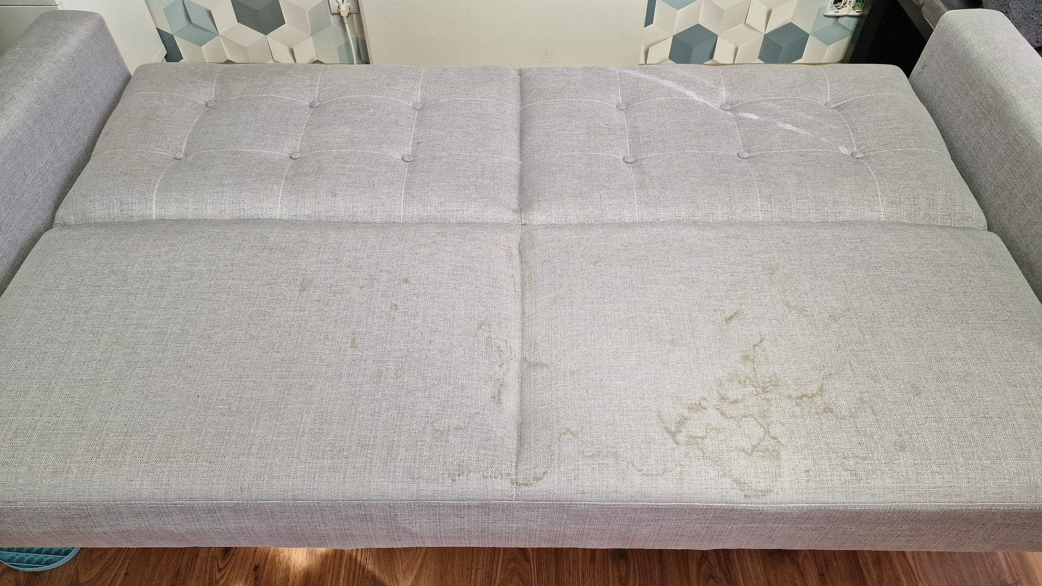 Szara rozkładana kanapa z widocznymi plamami przed czyszczeniem, widok z góry na siedzisko z guzikami, drewniana podłoga w tle.