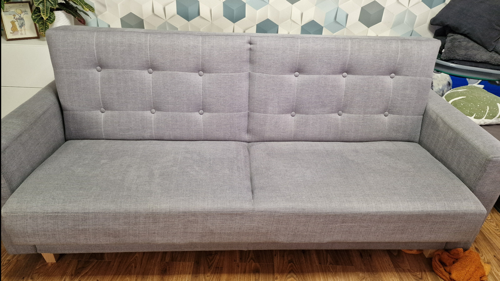 Szara, dwuosobowa kanapa z tapicerowanymi guzikami na oparciu, ustawiona na drewnianej podłodze z dekoracyjną ścianą w tle.