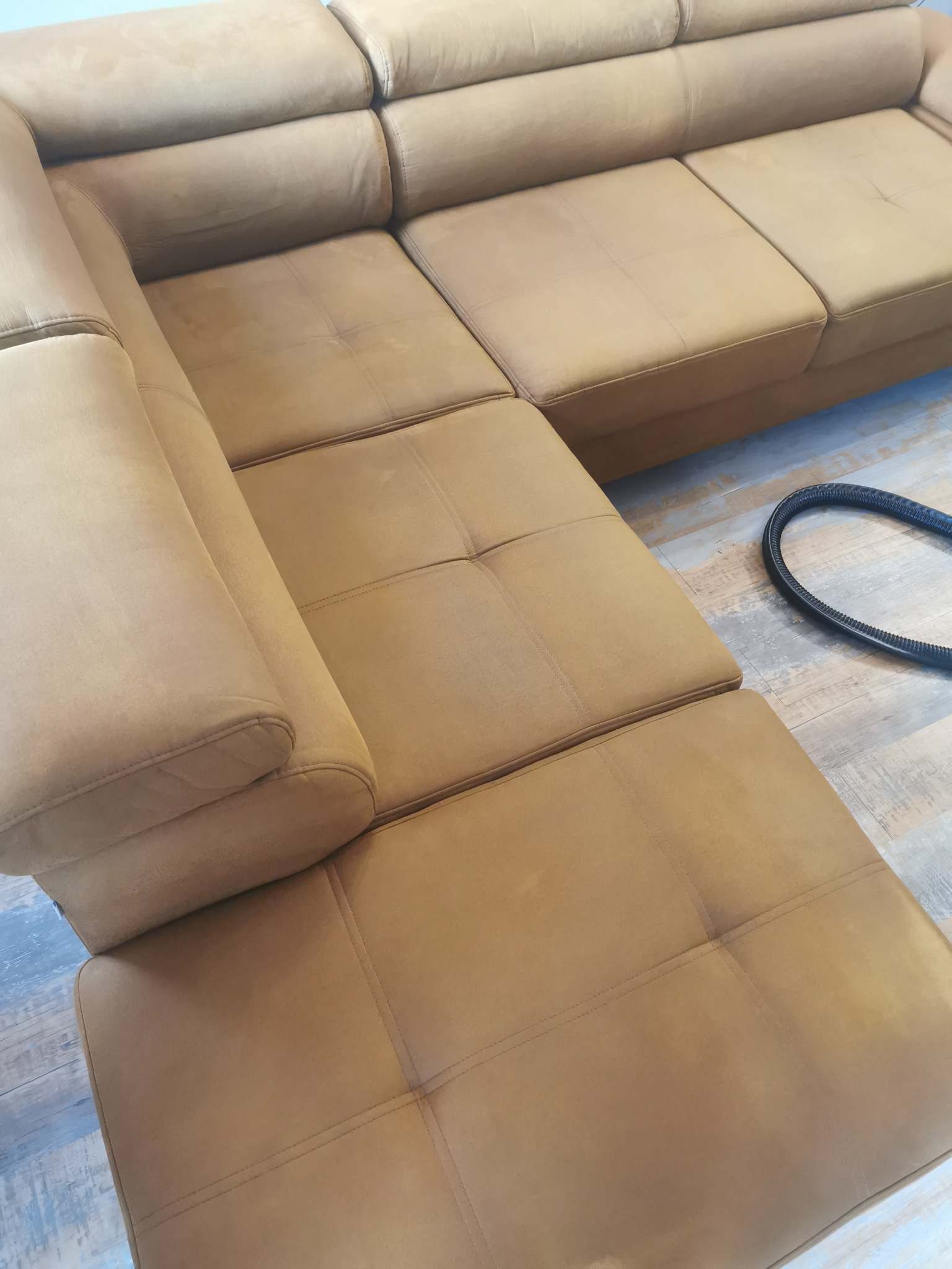 Jasnobrązowa, narożna kanapa z widocznym wężem od odkurzacza piorącego na podłodze z paneli.