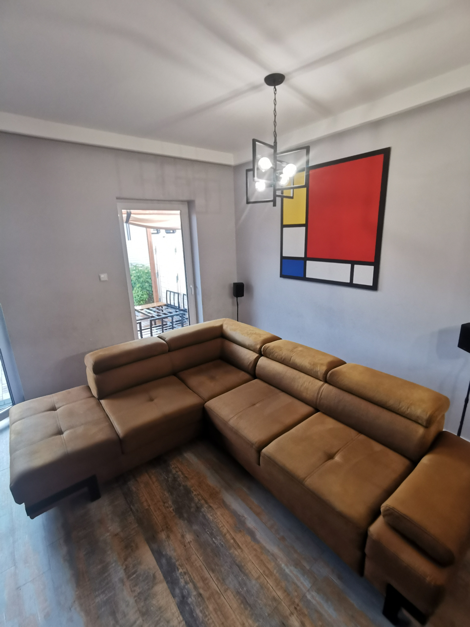 Brązowa, modułowa kanapa narożna z tapicerką w odcieniu musztardowym, ustawiona w salonie z szarymi ścianami i obrazem w stylu Mondriana, widoczna lampa sufitowa o geometrycznym kształcie.