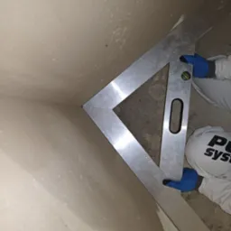 Pomiar kąta prostego w narożniku ściany po nałożeniu tynku, z użyciem aluminiowej kątownicy z poziomicą, pracownik w niebieskich rękawicach i białym kombinezonie.