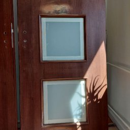 Perfect Tynk - Ciemnobrązowe drzwi z trzema szybami zabezpieczonymi taśmą malarską, widoczne uszkodzenia i ślady po naprawie na drewnianej ramie, cień rośliny na drzwiach.