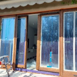 Perfect Tynk - Drewniane okno balkonowe zabezpieczone folią malarską przed renowacją. Widoczna drabina malarska i fragment wnętrza domu. Ujęcie z zewnątrz, w słoneczny dzień.