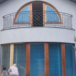 Perfect Tynk - Pracownik w kombinezonie przygotowuje drewniane okna do malowania, zabezpieczone folią, na tle szarej elewacji domu z balkonem i półokrągłym oknem.