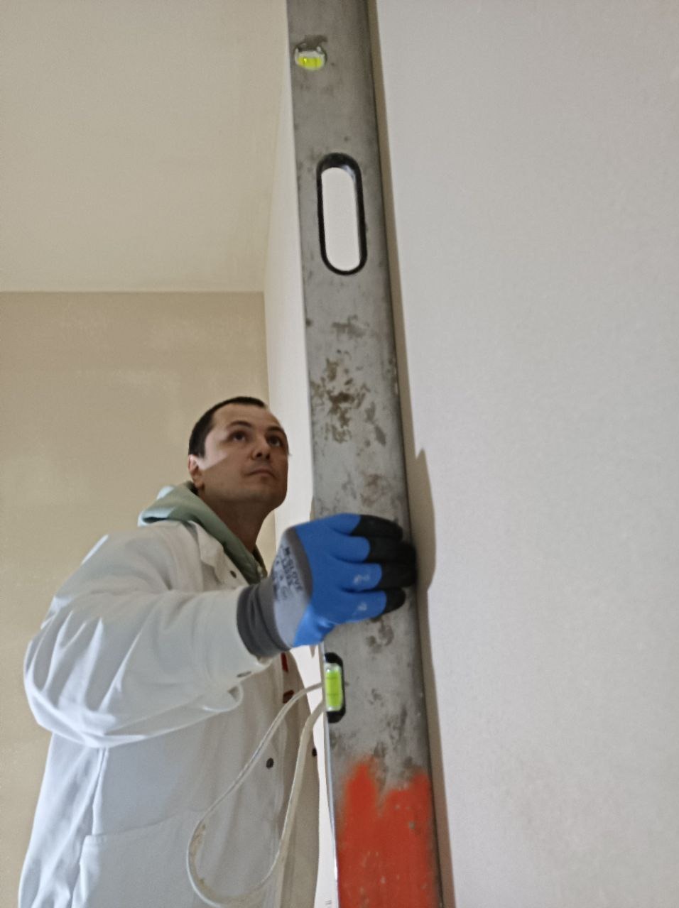 Pracownik w białym kombinezonie, z niebieskimi rękawicami, sprawdza pion za pomocą poziomicy na świeżo położonym tynku na ścianie.