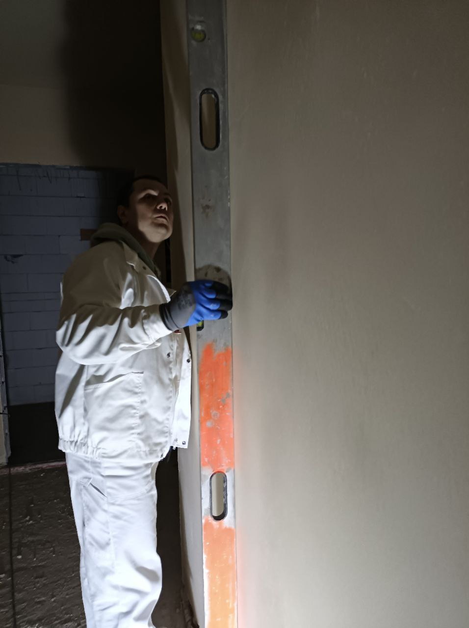 Pracownik w białym kombinezonie i niebieskich rękawicach, sprawdzający pionowość świeżo nałożonego tynku za pomocą poziomicy z pomarańczowymi akcentami.