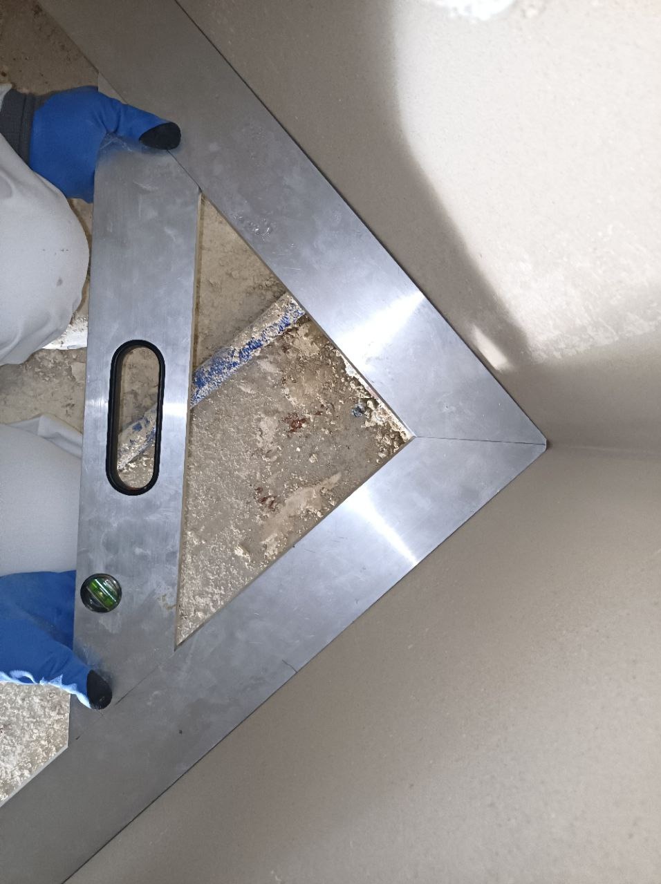 Metalowa łata kątowa z poziomicą oparta o świeżo nałożony tynk maszynowy przy ścianie, widoczne fragmenty ubrania roboczego i rękawic ochronnych.