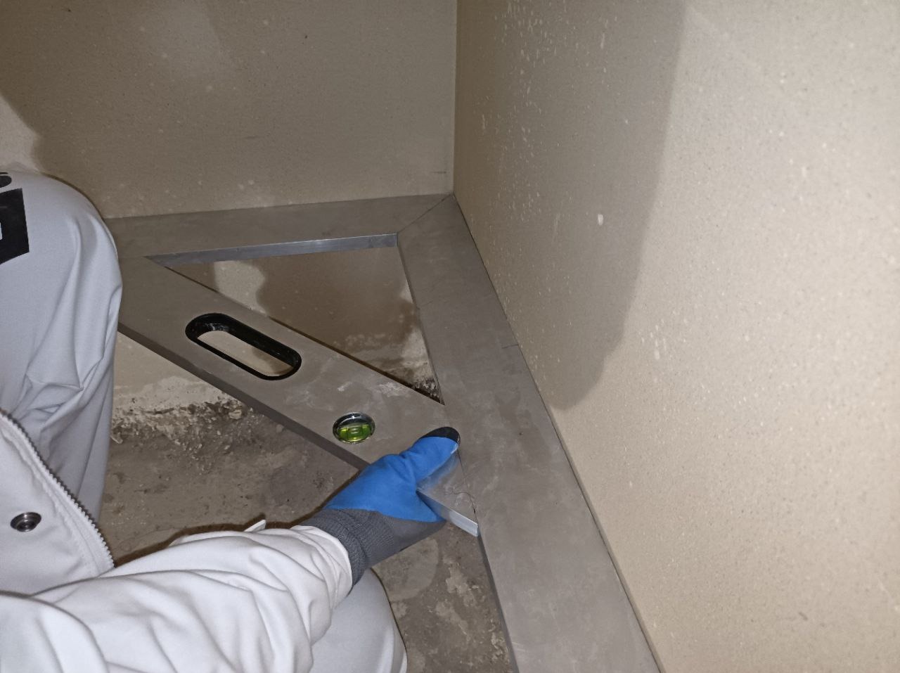 Pracownik w białym kombinezonie i niebieskich rękawicach sprawdza poziomicą świeżo nałożony tynk maszynowy w rogu pomieszczenia, widoczne niedoskonałości podłoża.