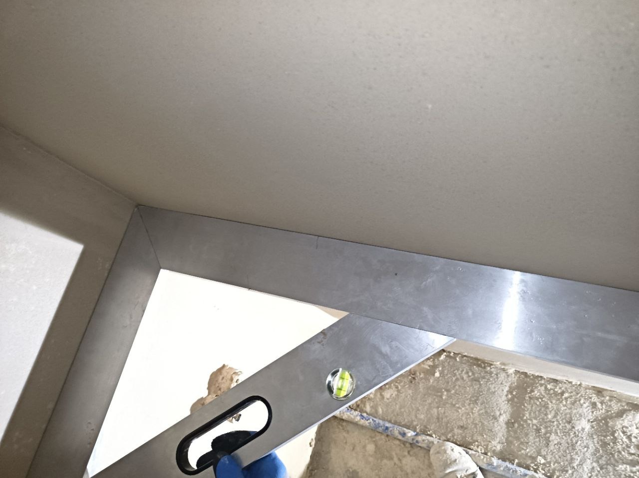 Świeżo położony tynk na suficie, sprawdzany poziomnicą w rogu pomieszczenia, widoczny fragment podłogi z resztkami zaprawy.