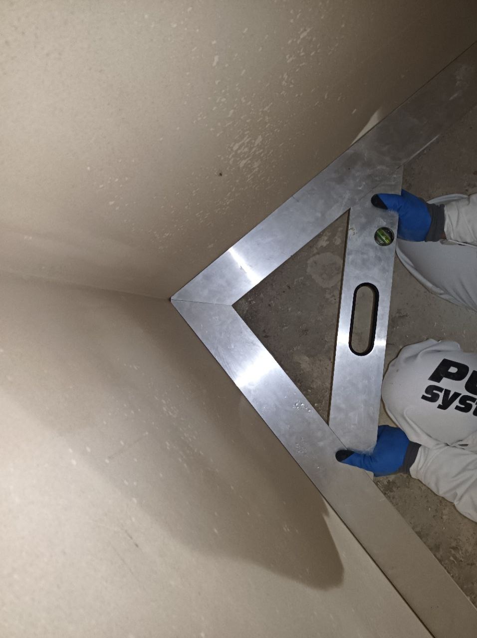 Pomiar kąta prostego w narożniku ściany po nałożeniu tynku, z użyciem aluminiowej kątownicy z poziomicą, pracownik w niebieskich rękawicach i białym kombinezonie.