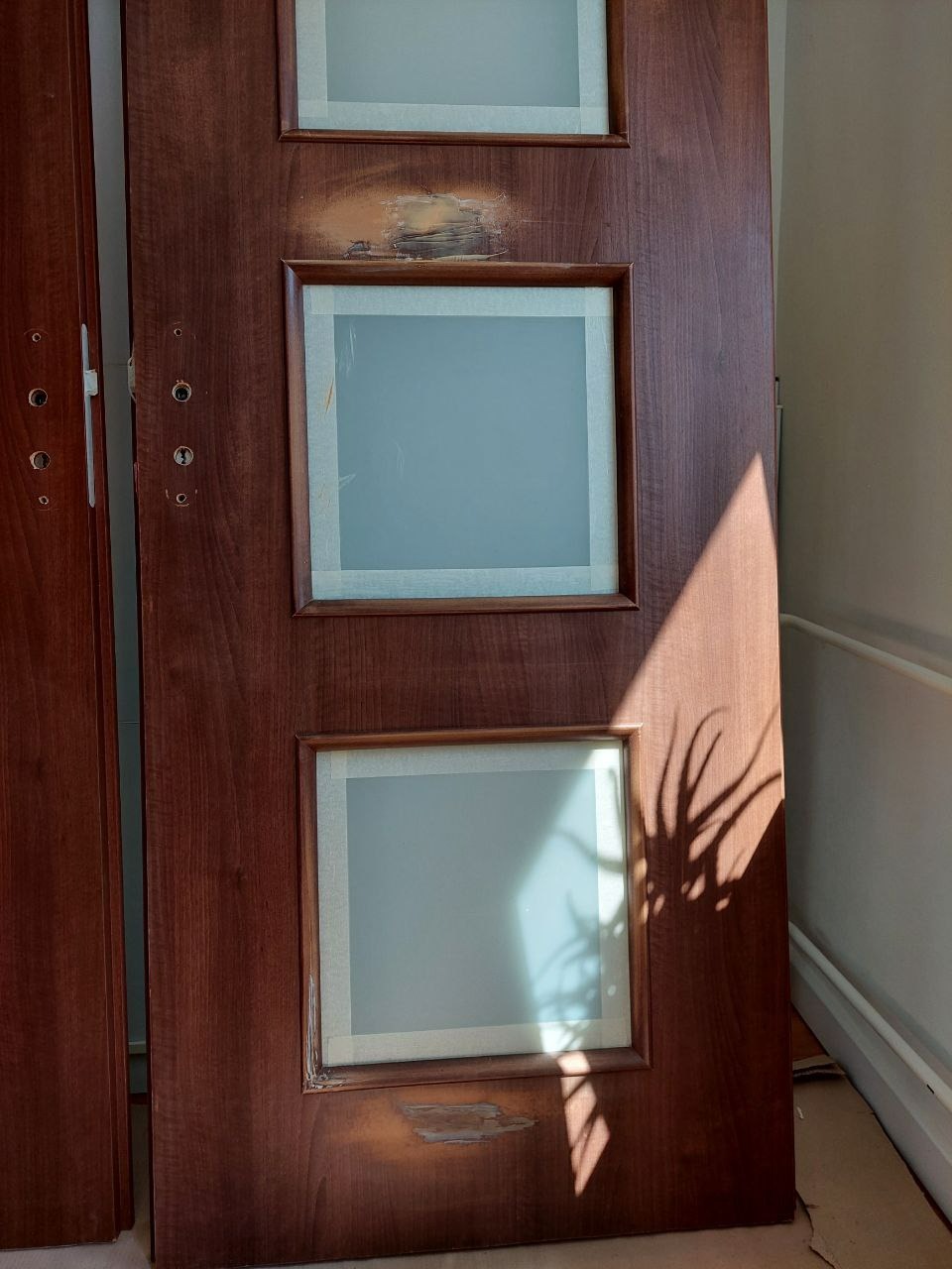 Ciemnobrązowe drzwi z trzema szybami zabezpieczonymi taśmą malarską, widoczne uszkodzenia i ślady po naprawie na drewnianej ramie, cień rośliny na drzwiach.