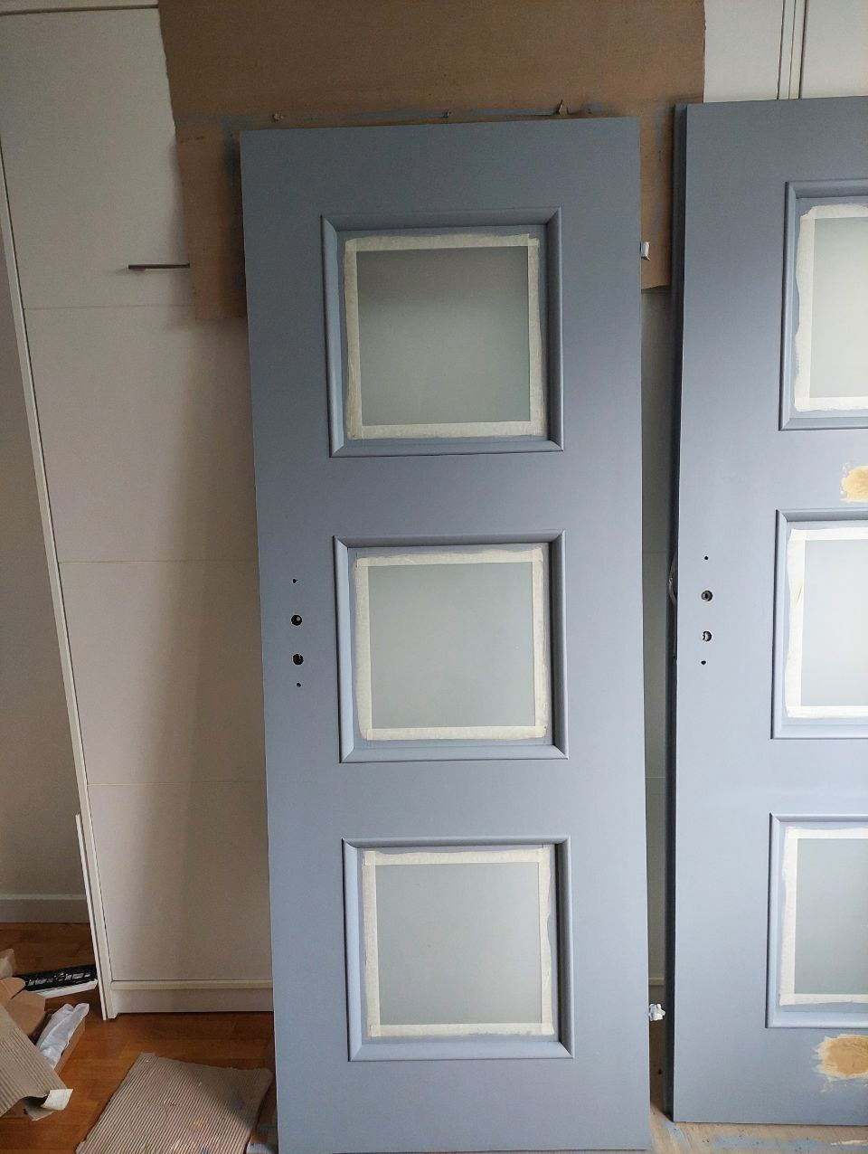 Dwie szare, odnowione drzwi z trzema kwadratowymi przeszkleniami, zabezpieczonymi taśmą malarską. Widoczne ślady szpachli i przygotowania do malowania.