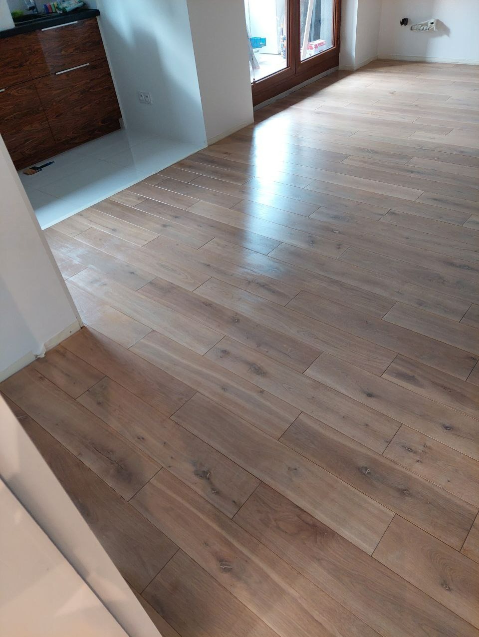 Ułożona podłoga z paneli drewnopodobnych w jasnym wnętrzu. Widoczne fragmenty ścian i szafki. Naturalne światło podkreśla strukturę drewna.