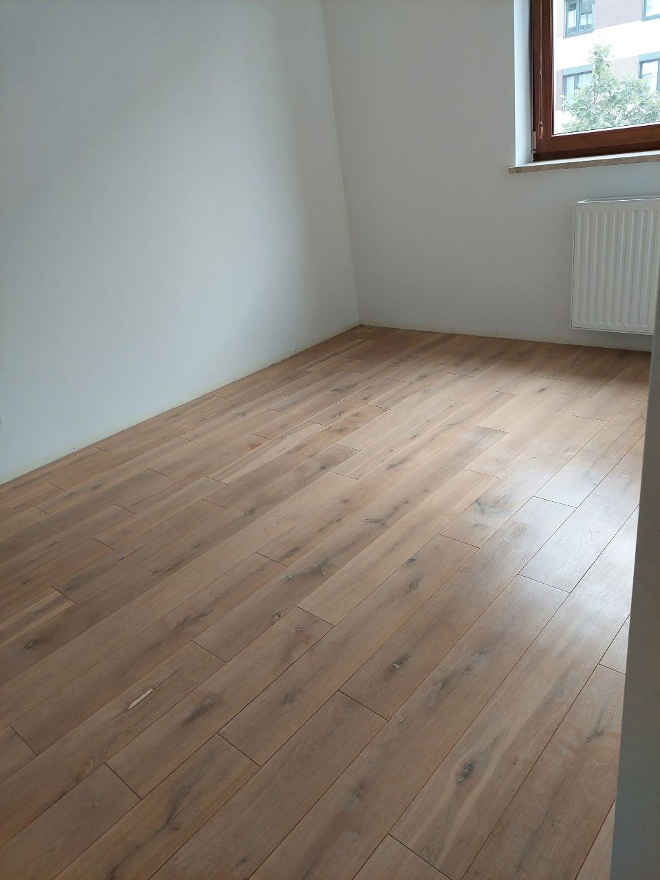 Jasne, drewniane panele podłogowe ułożone w pokoju z białymi ścianami i oknem. Widoczny fragment grzejnika. Podłoga po cyklinowaniu lub nowa.