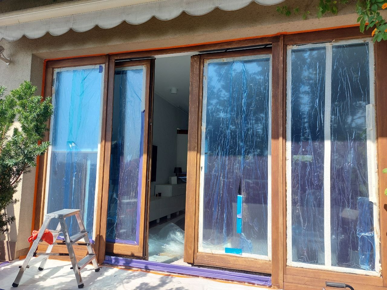 Drewniane okno balkonowe zabezpieczone folią malarską przed renowacją. Widoczna drabina malarska i fragment wnętrza domu. Ujęcie z zewnątrz, w słoneczny dzień.