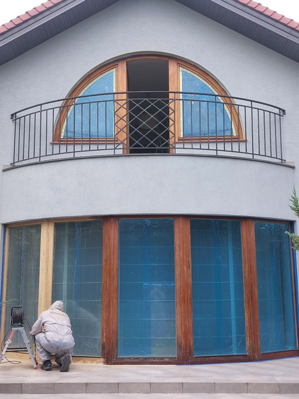 Pracownik w kombinezonie przygotowuje drewniane okna do malowania, zabezpieczone folią, na tle szarej elewacji domu z balkonem i półokrągłym oknem.
