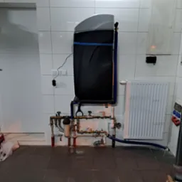 Filtr do wody zamontowany na ścianie z rurami miedzianymi i niebieskim wężem odprowadzającym wodę, widoczny grzejnik żeberkowy i białe płytki ścienne.