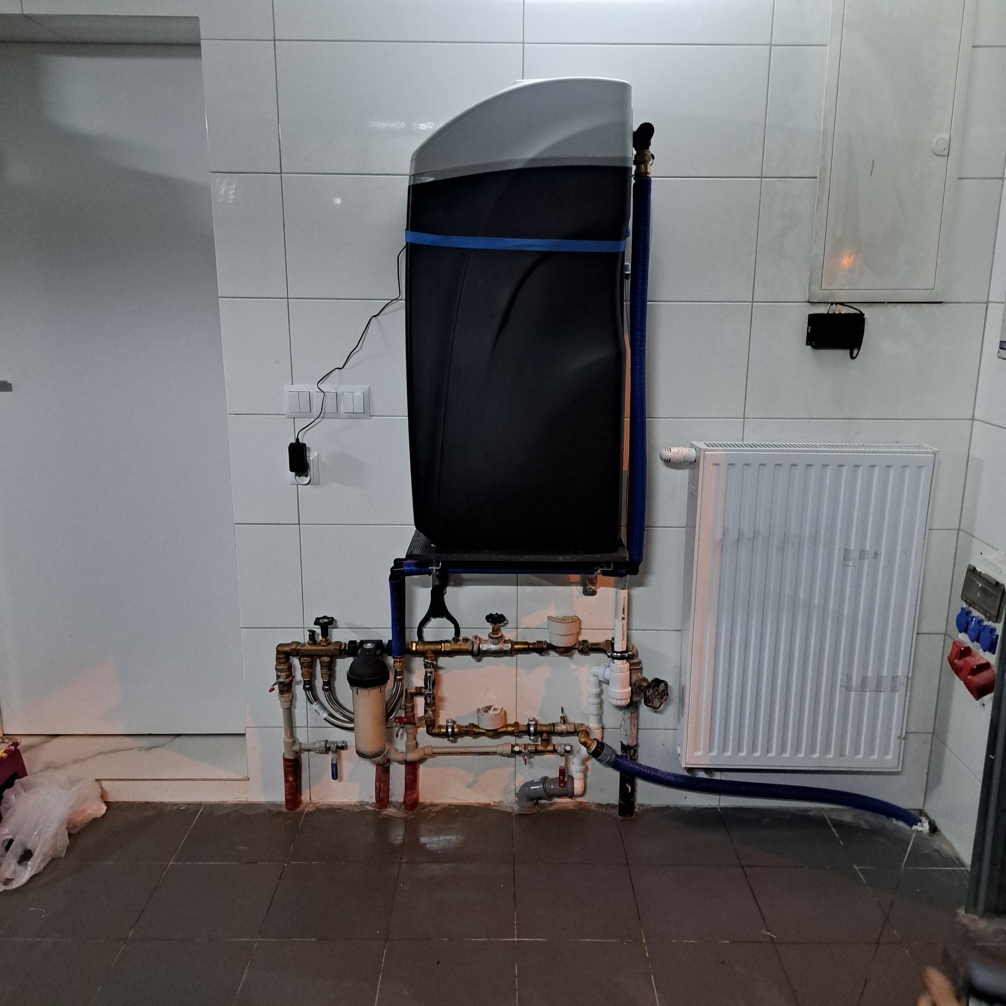 Filtr do wody zamontowany na ścianie z rurami miedzianymi i niebieskim wężem odprowadzającym wodę, widoczny grzejnik żeberkowy i białe płytki ścienne.