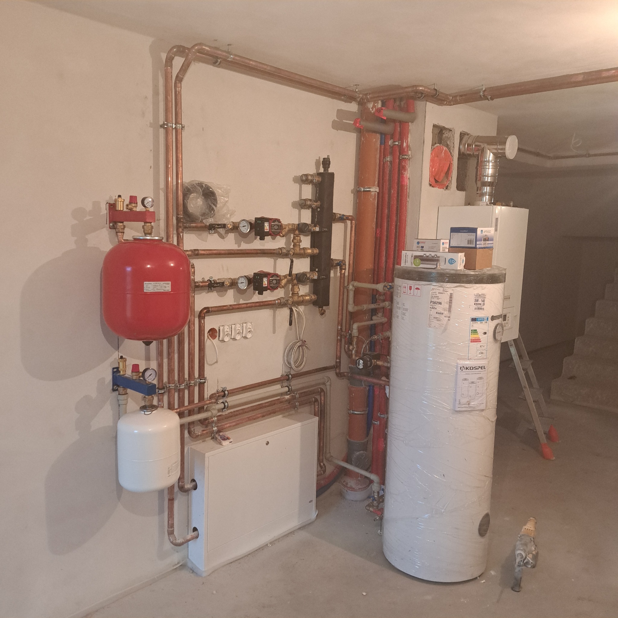 Nowa instalacja grzewcza z kotłem, zasobnikiem owiniętym folią, czerwonym naczyniem przeponowym, licznymi miedzianymi rurami i zaworami, zamontowana w surowym pomieszczeniu.