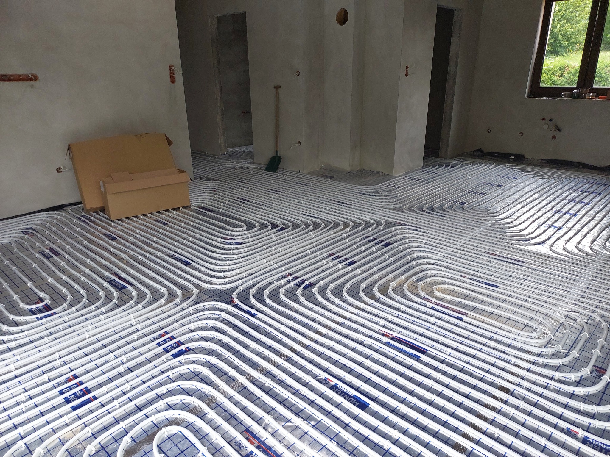 Rozłożone białe rury ogrzewania podłogowego na siatce w surowym wnętrzu, widoczne kartony i łopata, przygotowane do zalania wylewką.