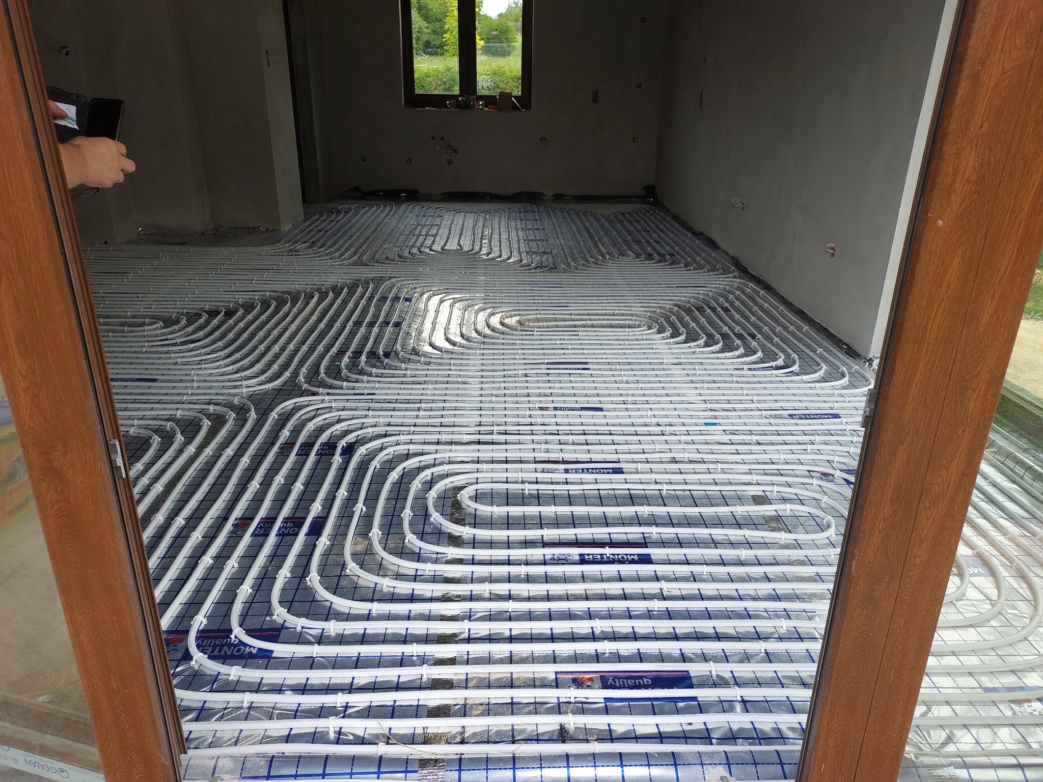 Białe rury ogrzewania podłogowego ułożone w pętle na folii z nadrukowaną siatką w surowym pomieszczeniu, widok zza drewnianej ramy okiennej.