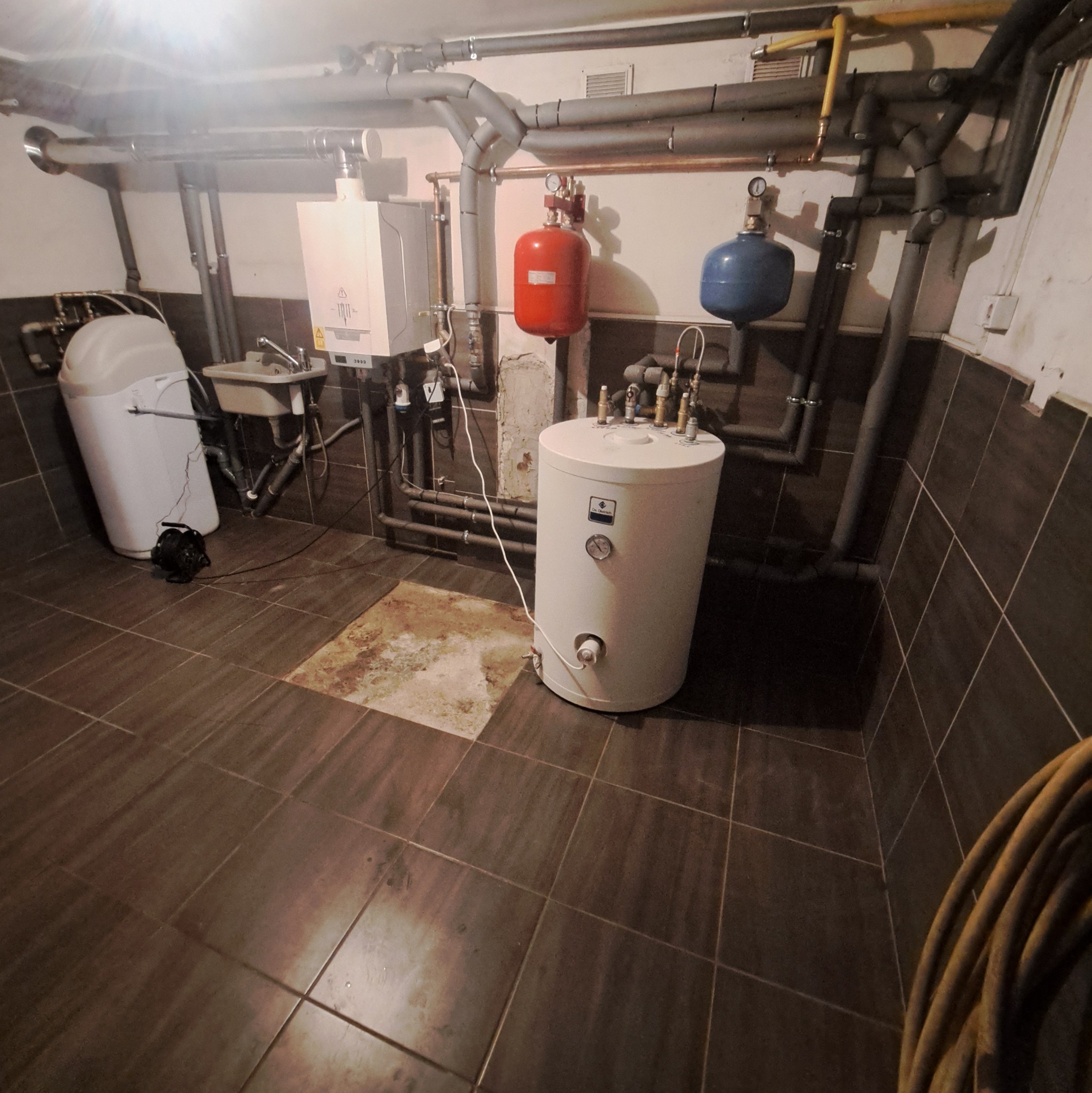 Kotłownia z piecem gazowym, zasobnikiem ciepłej wody, zmiękczaczem wody i rozbudowaną instalacją hydrauliczną z rurami w izolacji, widoczne manometry i zawory.