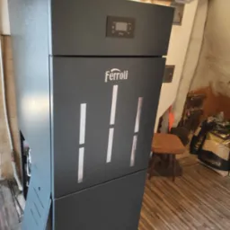 Szary piec CO marki Ferroli z widocznym wyświetlaczem i workiem ekopelletu w tle, ustawiony w pomieszczeniu z drewnianą podłogą i widocznymi śladami wilgoci na suficie.