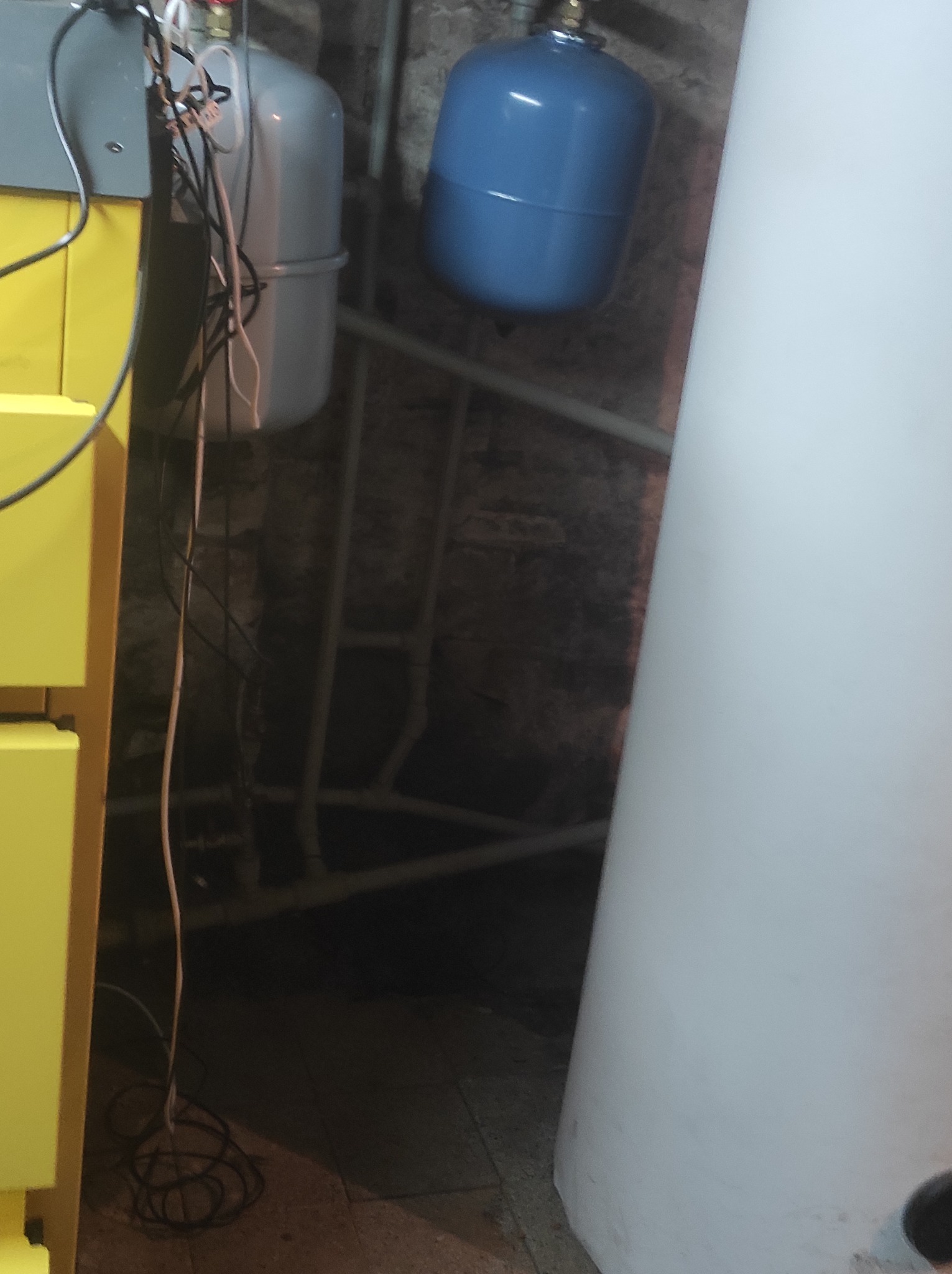 Fragment kotłowni z żółtym piecem, szarym i niebieskim zbiornikiem wyrównawczym na tle ceglanej ściany i instalacji rurowej.