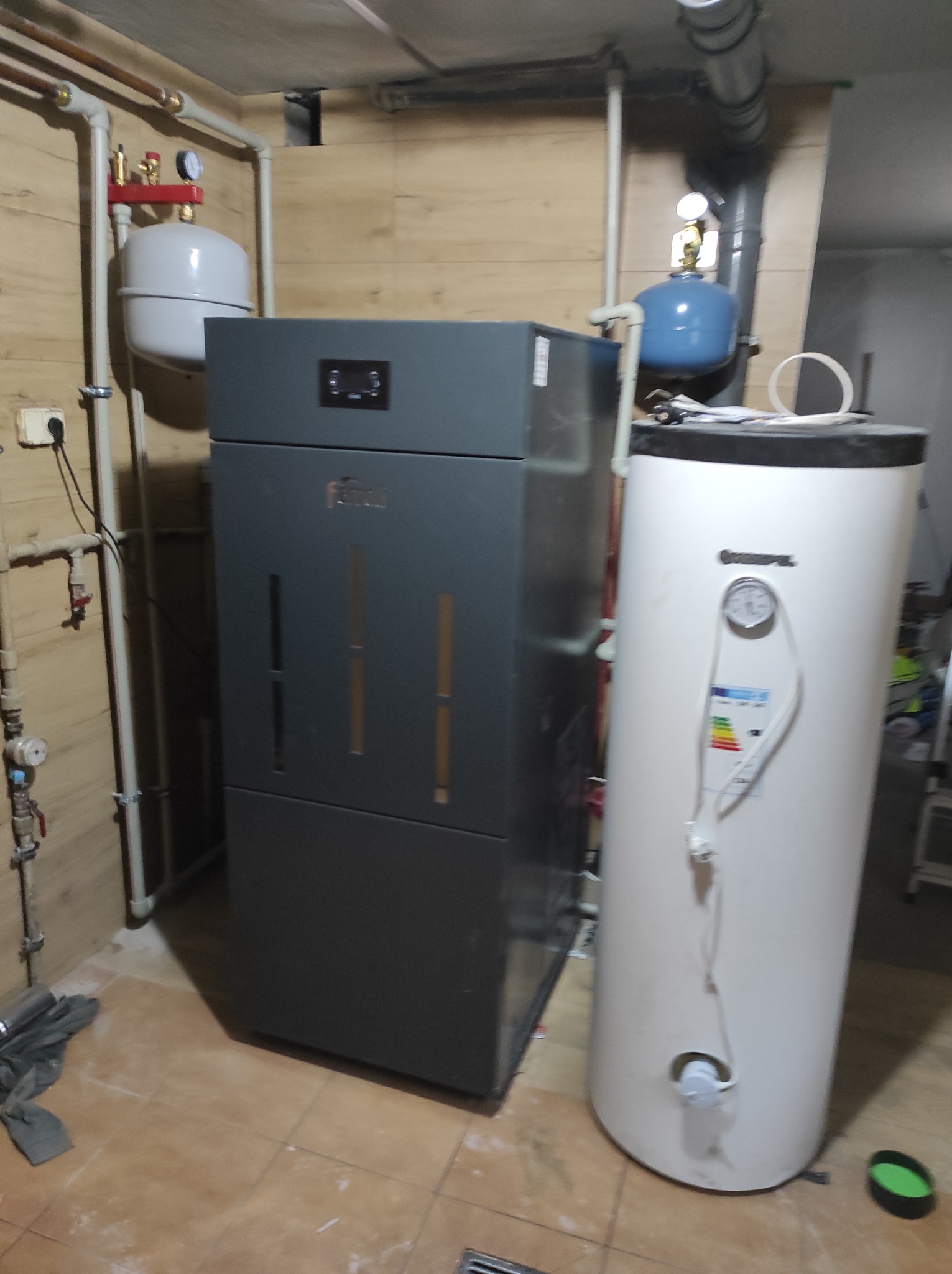 Szary piec centralnego ogrzewania marki Ferroli obok białego zasobnika ciepłej wody z widocznym wskaźnikiem temperatury, instalacja w pomieszczeniu z wykończeniem w boazerii.