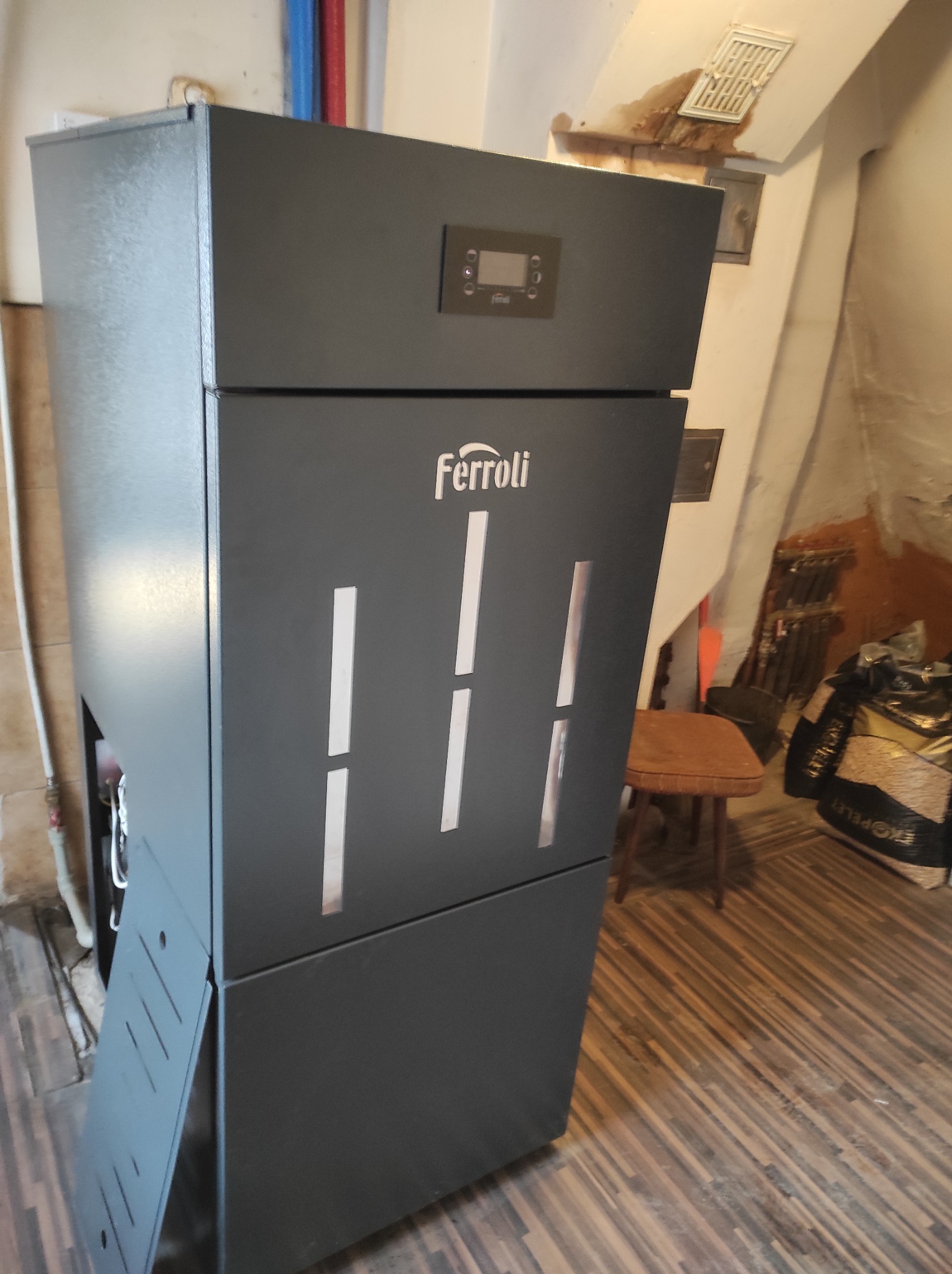 Szary piec CO marki Ferroli z widocznym wyświetlaczem i workiem ekopelletu w tle, ustawiony w pomieszczeniu z drewnianą podłogą i widocznymi śladami wilgoci na suficie.