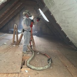 Bramex - Pracownik w kombinezonie ochronnym natryskuje piankę izolacyjną na poddasze, widoczne drewniane elementy konstrukcji dachu i fragment okna.