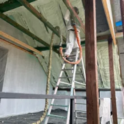 Pracownik w kombinezonie ochronnym na drabinie aplikuje piankę PUR na poddaszu, widoczne drewniane krokwie i izolacja z pianki.