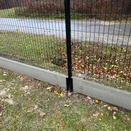 Bramex - Fragment czarnego ogrodzenia panelowego z podmurówką betonową, widok z bliska na słupek i przęsło, z widoczną linią wytyczającą przebieg ogrodzenia na trawie.