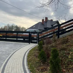 Nowoczesna, przesuwna brama wjazdowa z czarnymi metalowymi elementami i poziomymi panelami z drewna, widoczna z zewnątrz posesji na tle domu z ciemną dachówką i podjazdem z kostki brukowej.