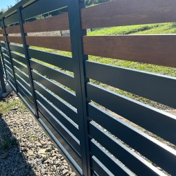 Bramex - Nowoczesne ogrodzenie z poziomych, szarych paneli metalowych i wstawkami z drewna kompozytowego, widok z zewnątrz na tle zielonego trawnika i drzew, żwirowe podłoże u podstawy.