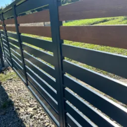 Nowoczesne ogrodzenie z poziomych, szarych paneli metalowych i wstawkami z drewna kompozytowego, widok z zewnątrz na tle zielonego trawnika i drzew, żwirowe podłoże u podstawy.