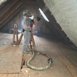 Bramex - Ocieplanie poddasza pianką PUR: pracownik w kombinezonie natryskuje pianę na skośny dach. Widoczna drabina i wąż do natrysku. Część poddasza już pokryta pianą.