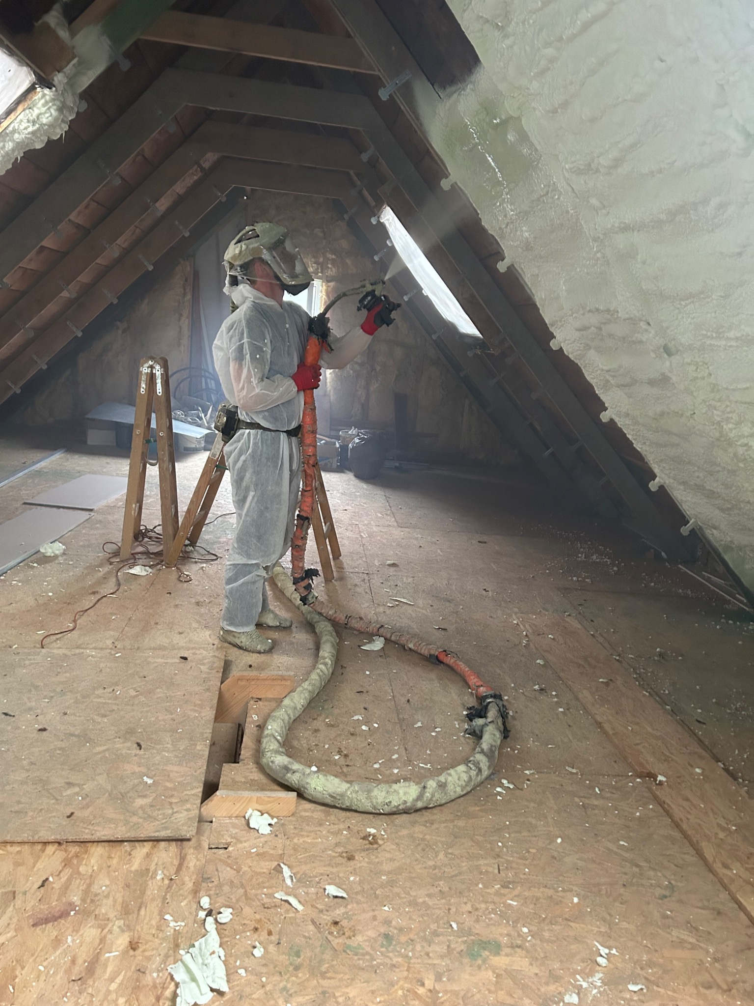 Pracownik w kombinezonie ochronnym natryskuje piankę izolacyjną na poddasze, widoczne drewniane elementy konstrukcji dachu i fragment okna.
