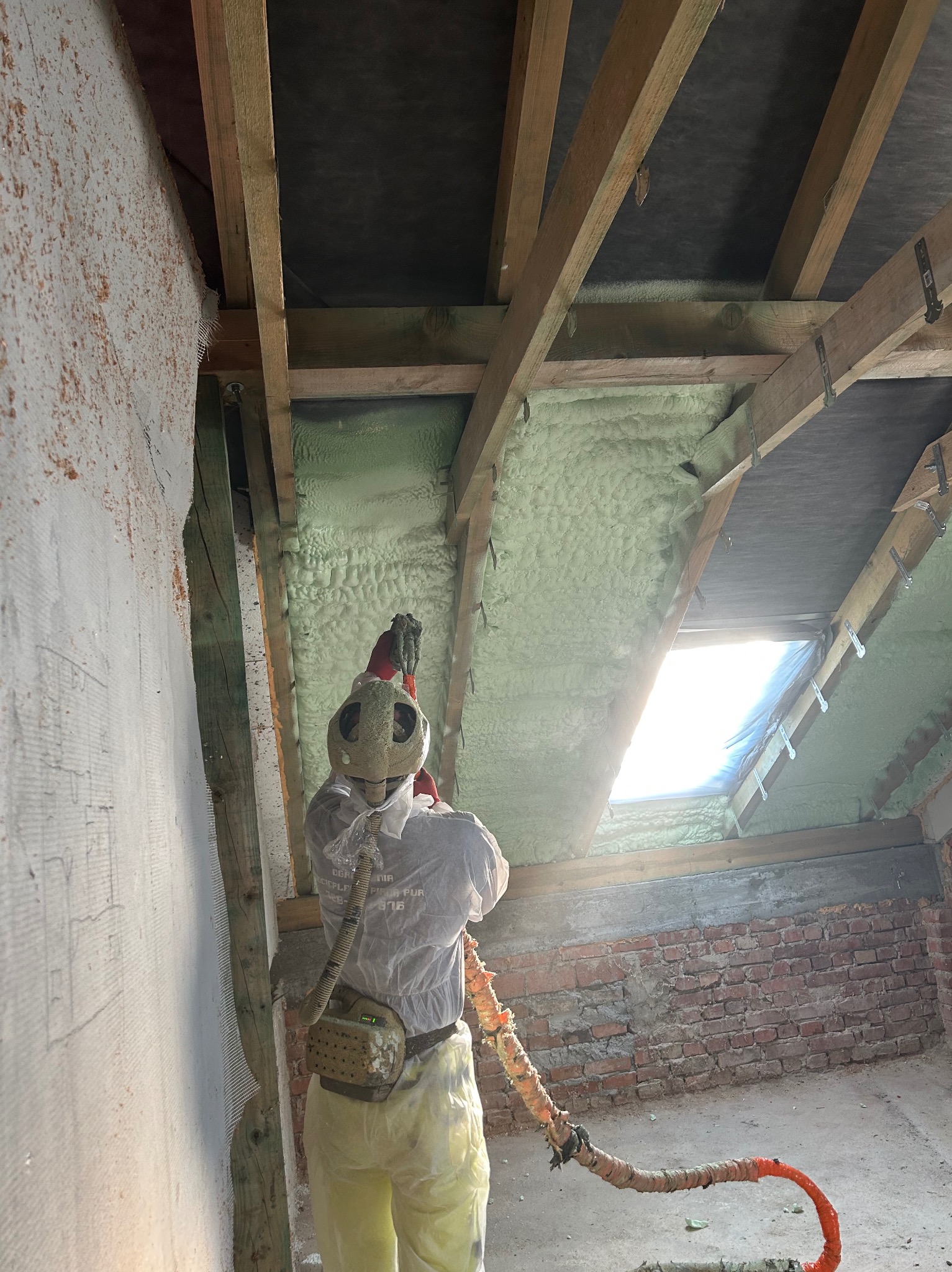Pracownik w kombinezonie ochronnym aplikuje piankę izolacyjną PUR na poddaszu, widoczne drewniane krokwie i fragment okna dachowego.