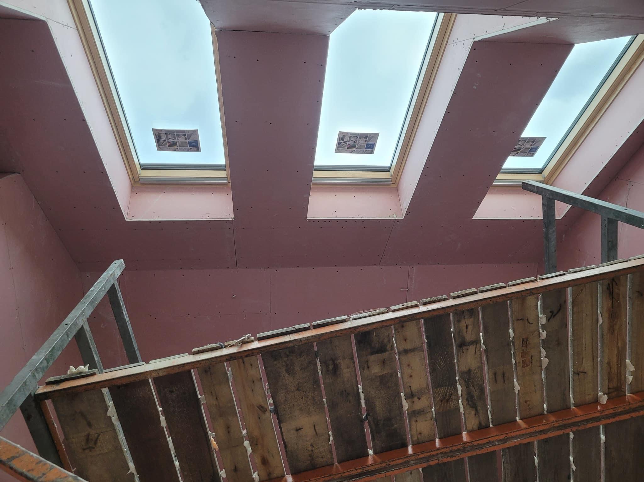 Wnętrze poddasza z zamontowanymi oknami dachowymi, wykończone płytami gipsowo-kartonowymi w kolorze różowym, widoczny fragment rusztowania.
