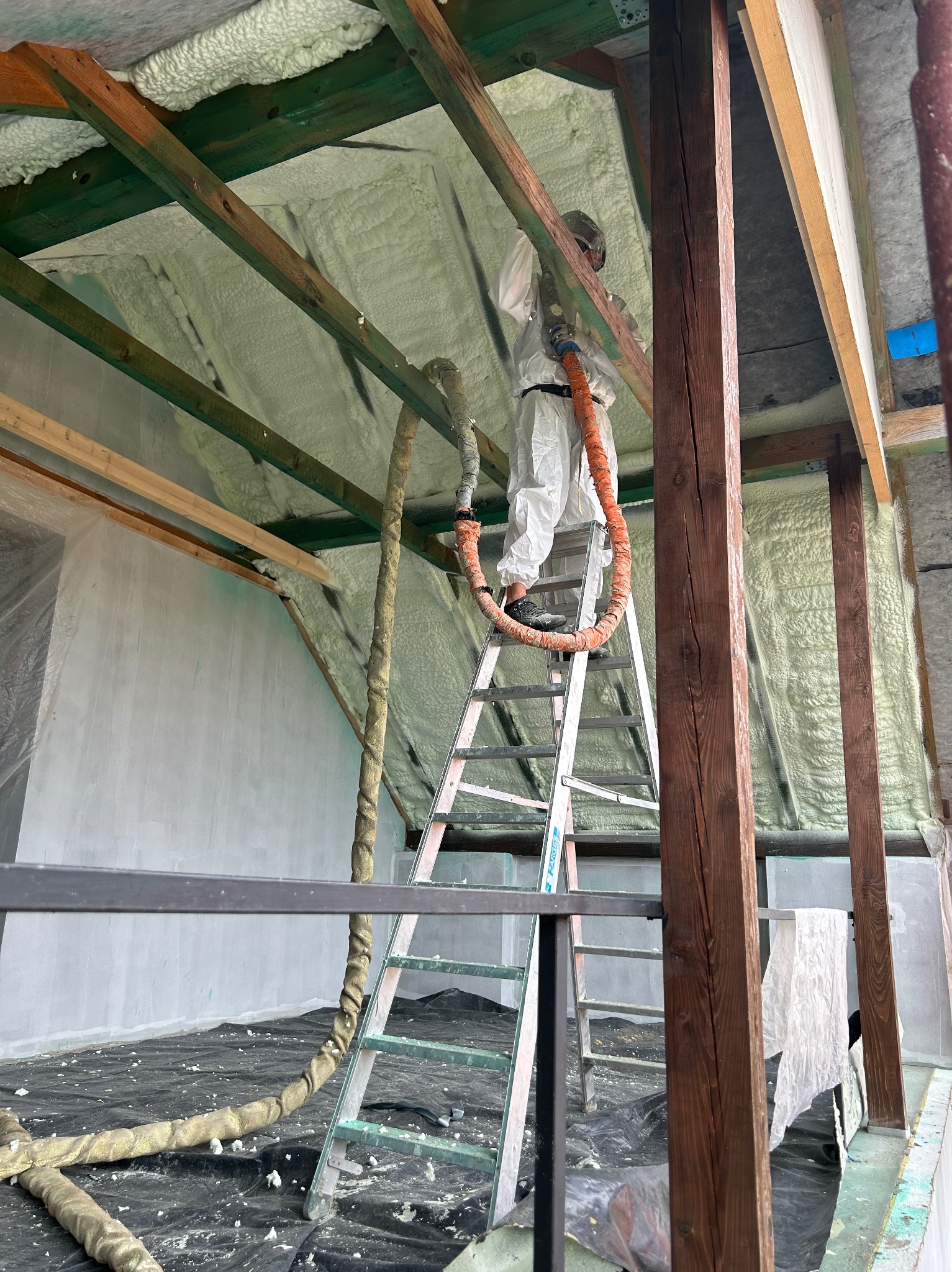 Pracownik w kombinezonie ochronnym na drabinie aplikuje piankę PUR na poddaszu, widoczne drewniane krokwie i izolacja z pianki.